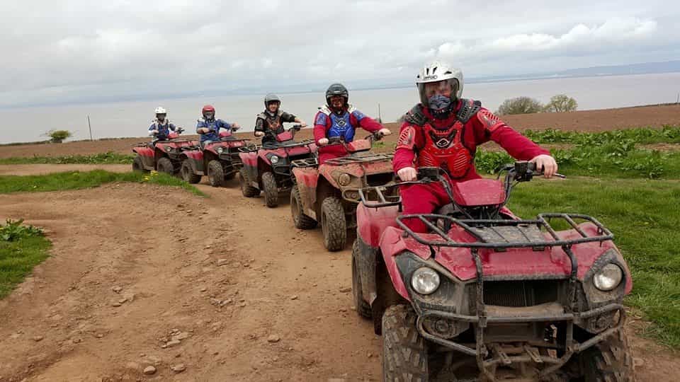 Five people wearing helmets and protective gear ride red quad bikes in a line along a dirt path on a cloudy day, with grassy fields and water visible in the background.