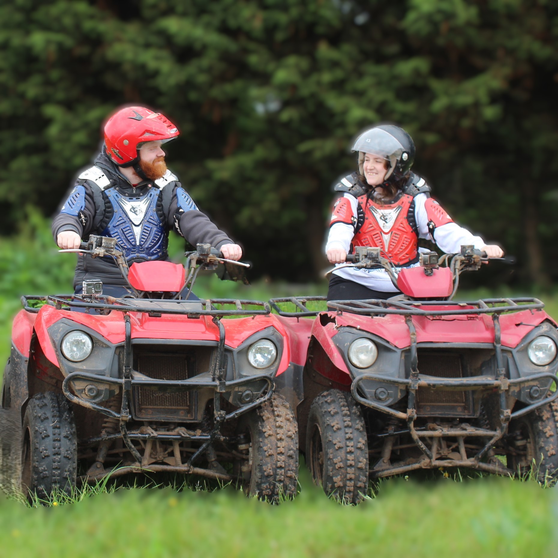 Two people in helmets and protective gear sit on muddy red ATVs facing each other and smiling, with green grass and blurred trees in the background.