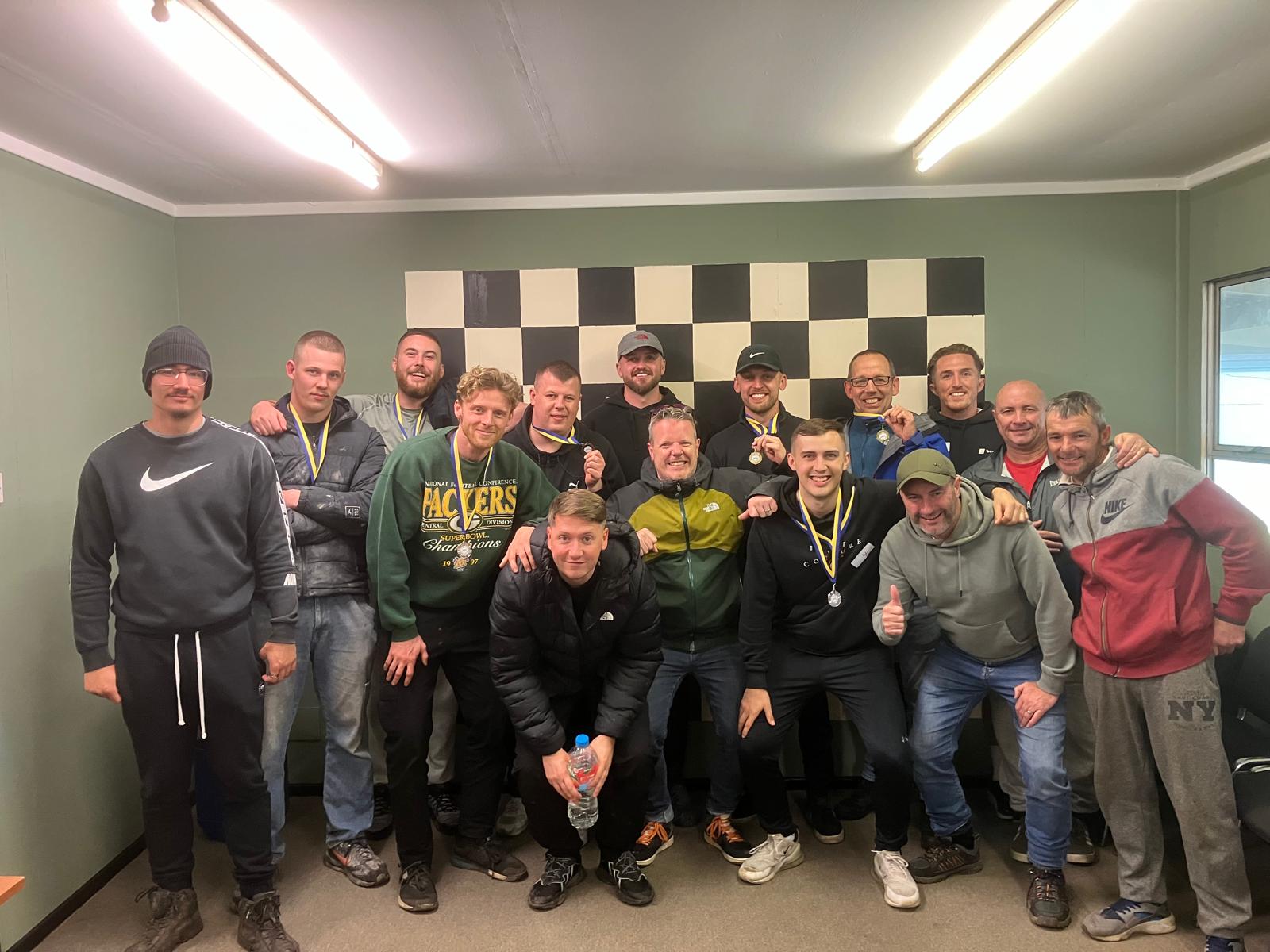 A group of sixteen men pose and smile indoors in front of a large black-and-white checkerboard. Some wear medals around their necks, and most are dressed in casual or sportswear. The atmosphere is celebratory and friendly.