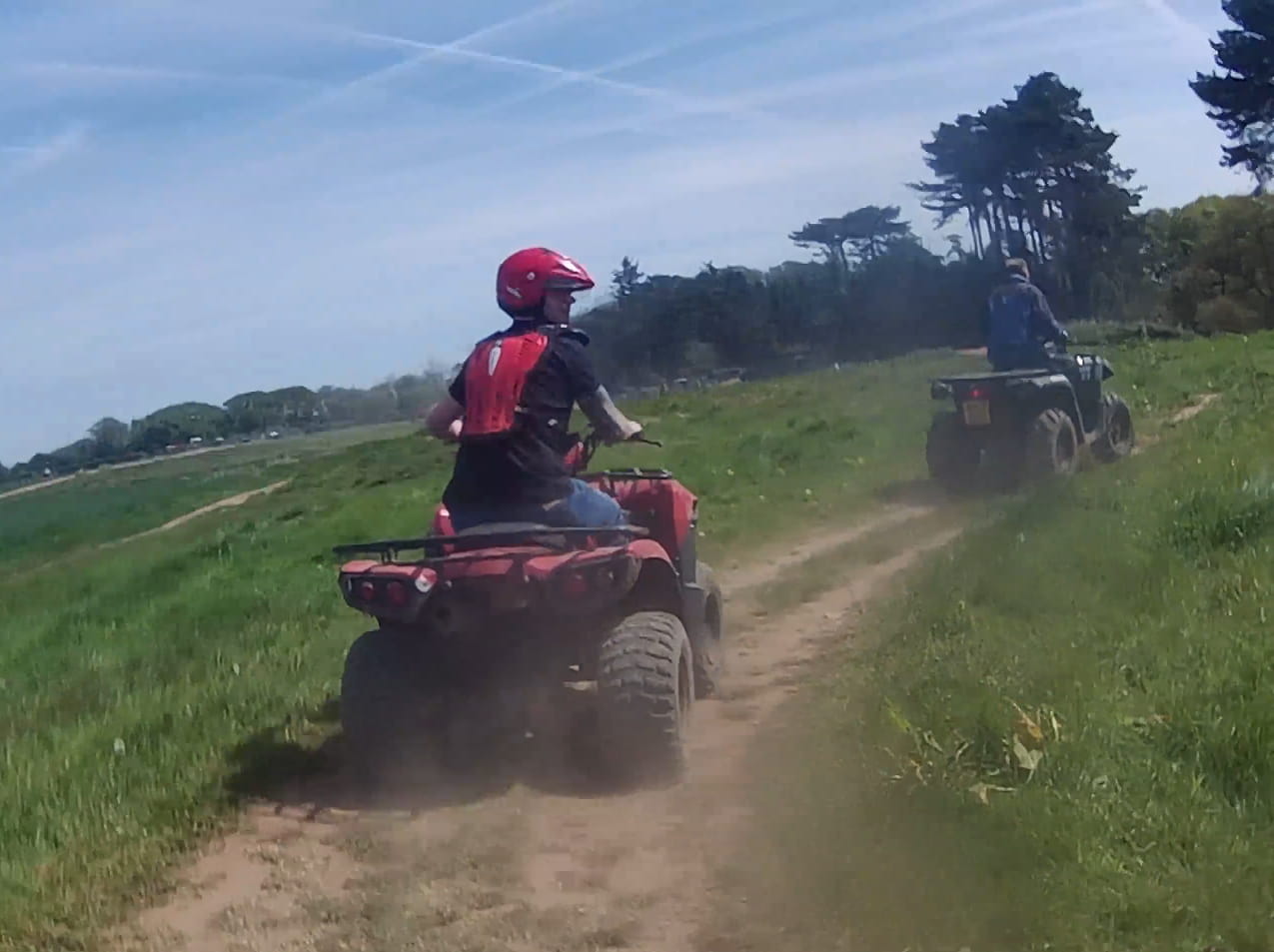 Two people ride ATVs on a dirt trail through a grassy field on a sunny day. One rider wears a red helmet and backpack, and the other rides ahead. Trees and blue sky are visible in the background.